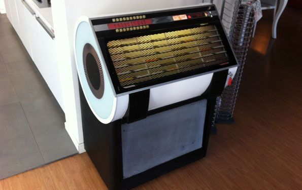 Jukebox bianco e nero con un grande altoparlante circolare. Appoggiato a una parete bianca e a un pavimento in legno.