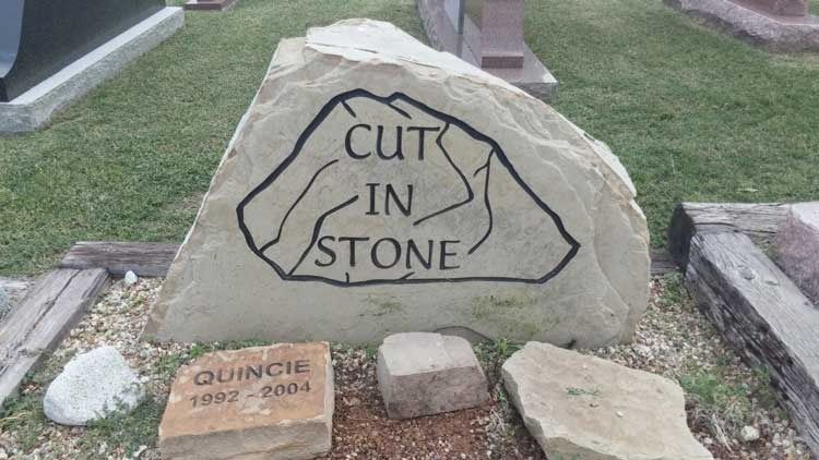 Stone monument engraved “CUT IN STONE” in a cemetery, with a small plaque and flowers at the base