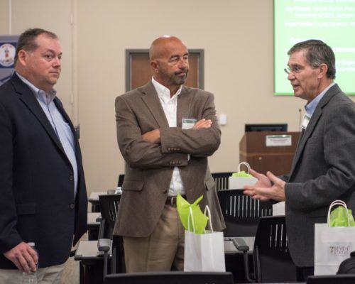 Three men in suits are standing in a classroom talking to each other