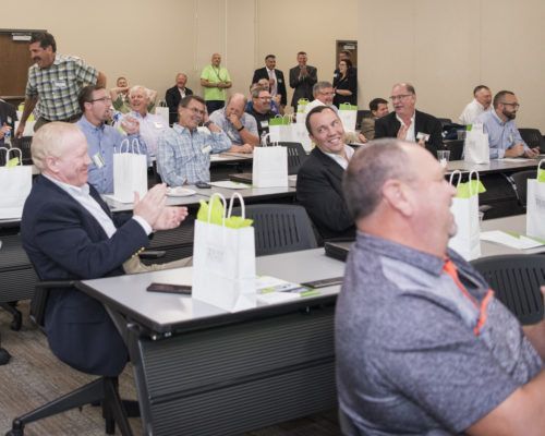 A group of men are sitting at tables in a classroom applauding.