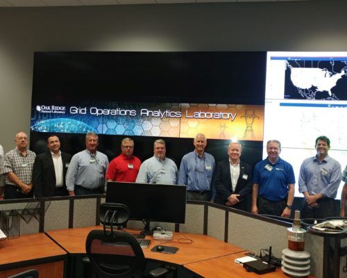 A group of men are standing in front of a large screen that says grid operations analytics laboratory