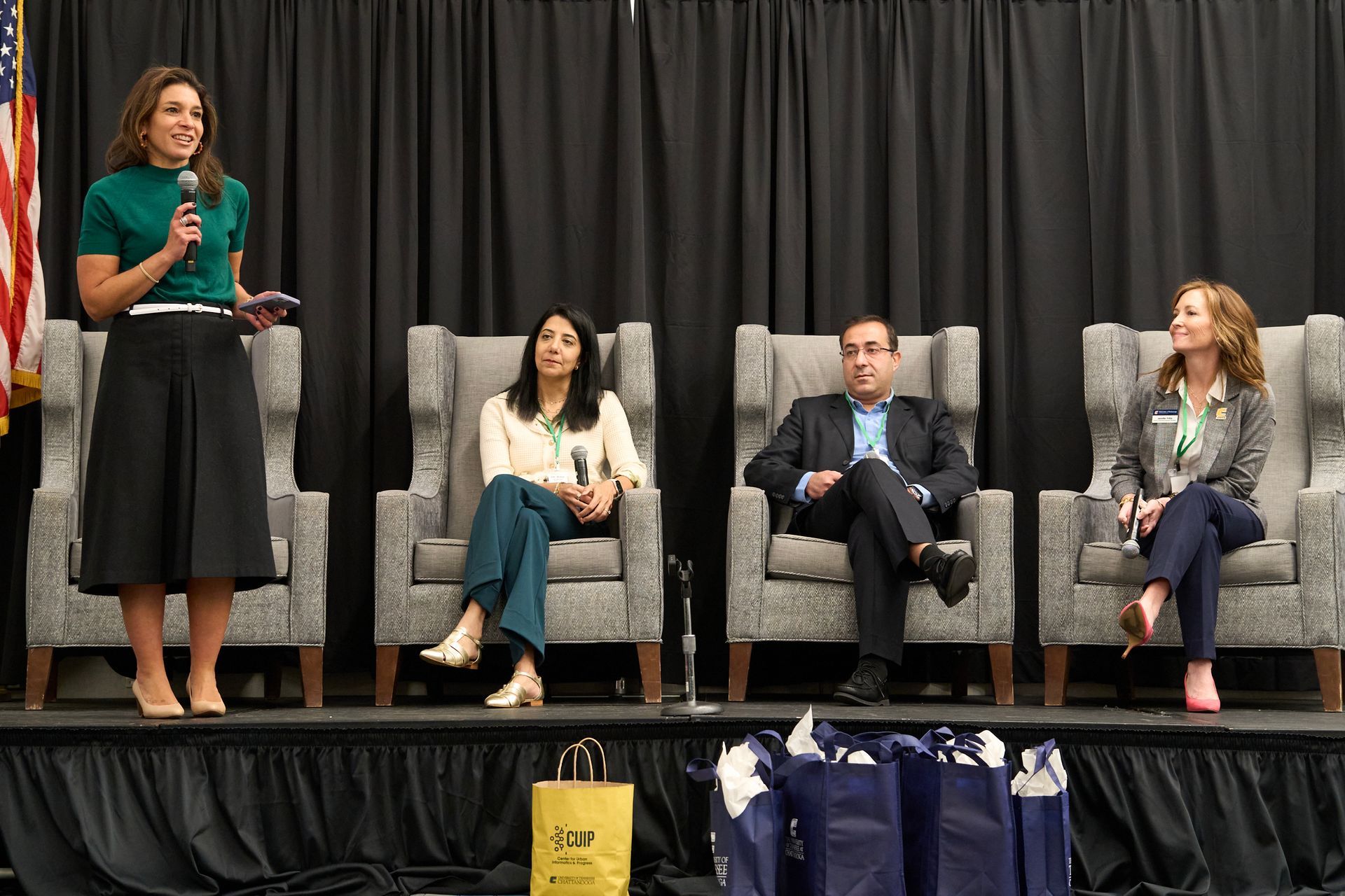 A woman is standing on a stage talking into a microphone while three people sit in chairs.
