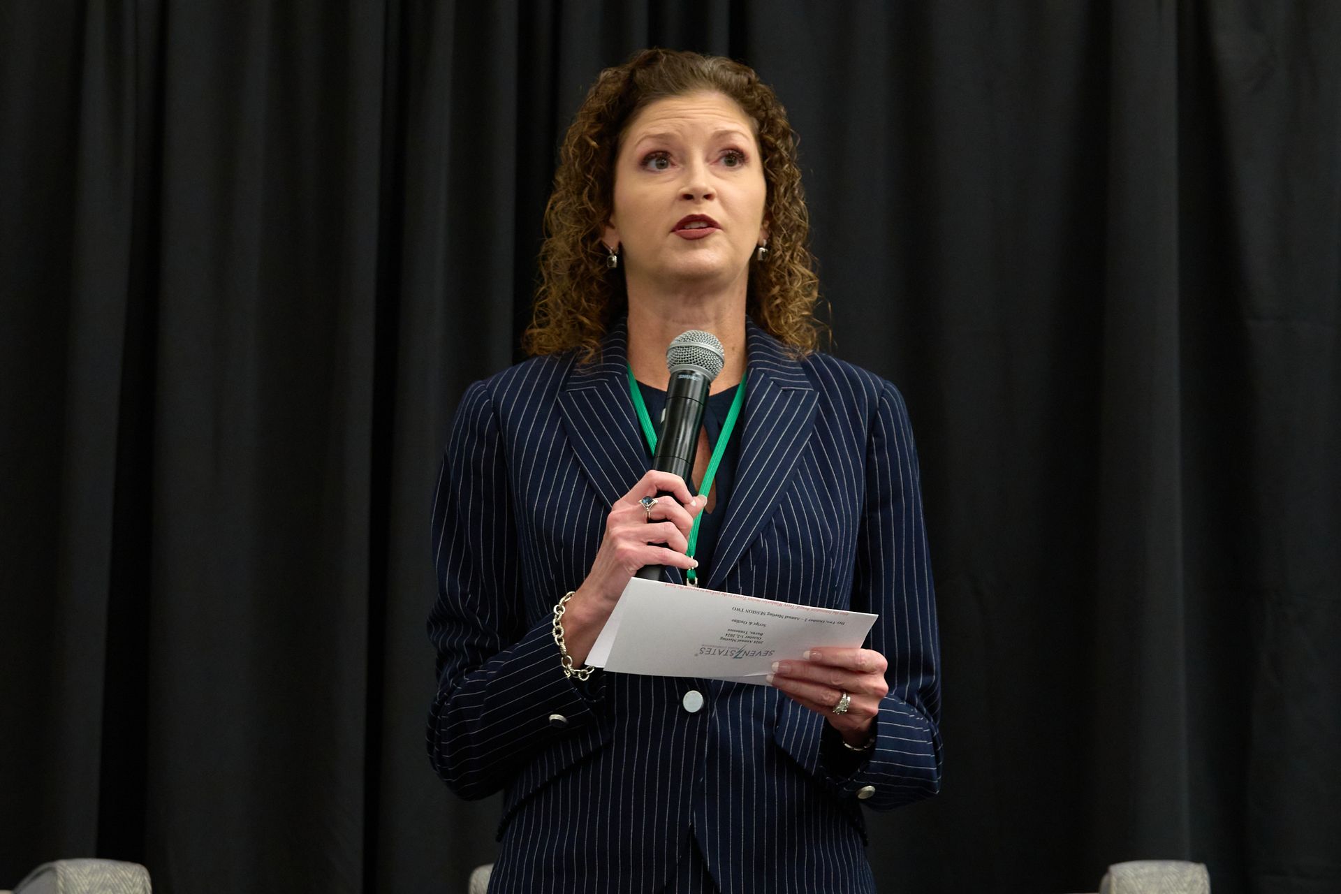 A woman is holding a microphone and a piece of paper in front of a black curtain.