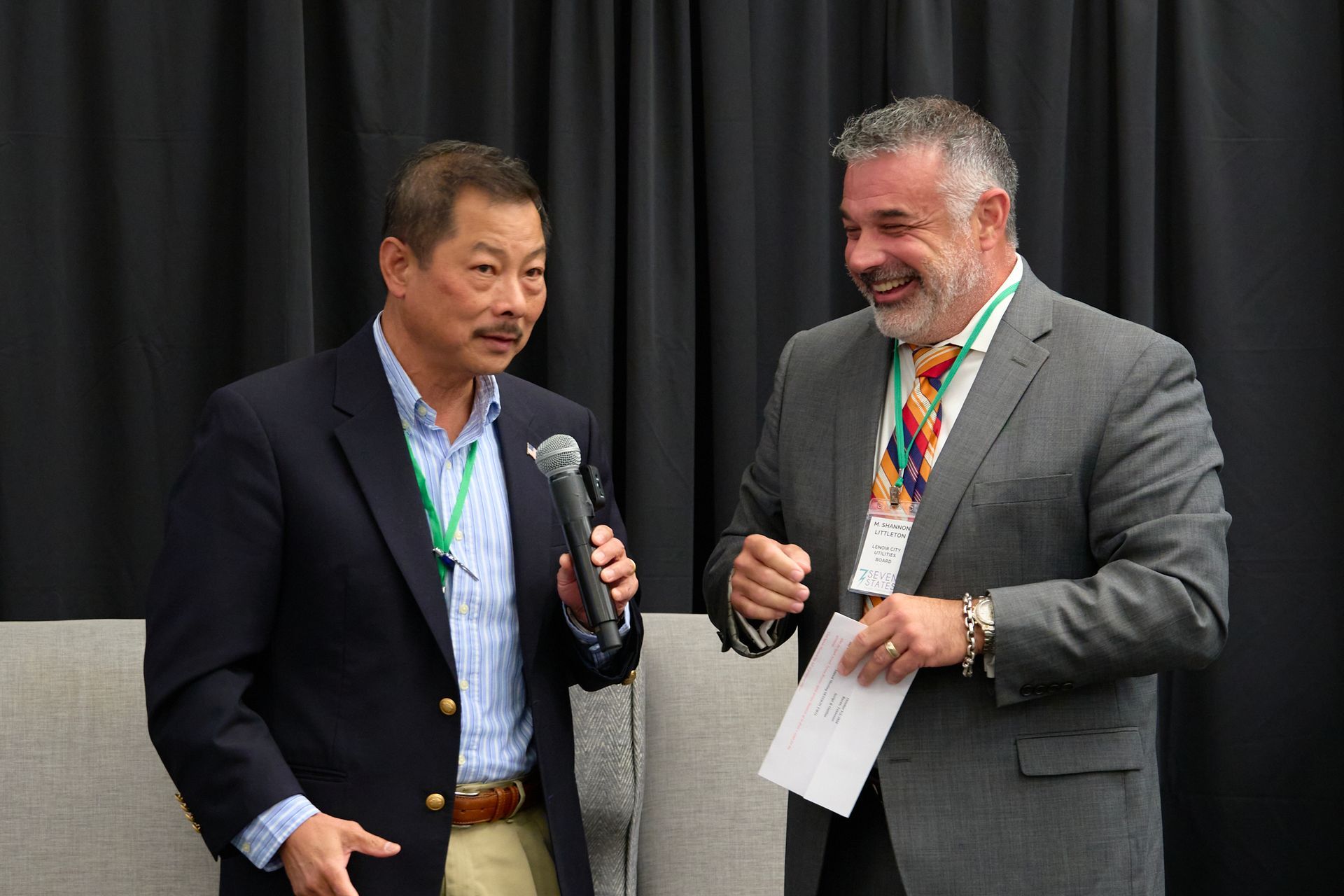 Two men in suits are standing next to each other talking into microphones.