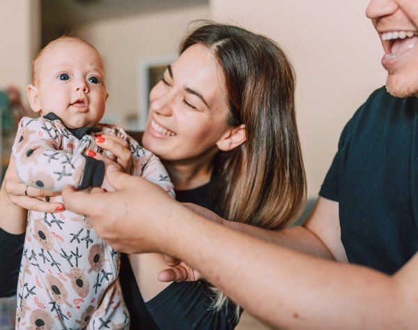 A woman and man smiling at a baby in floral onesie; indoor setting.