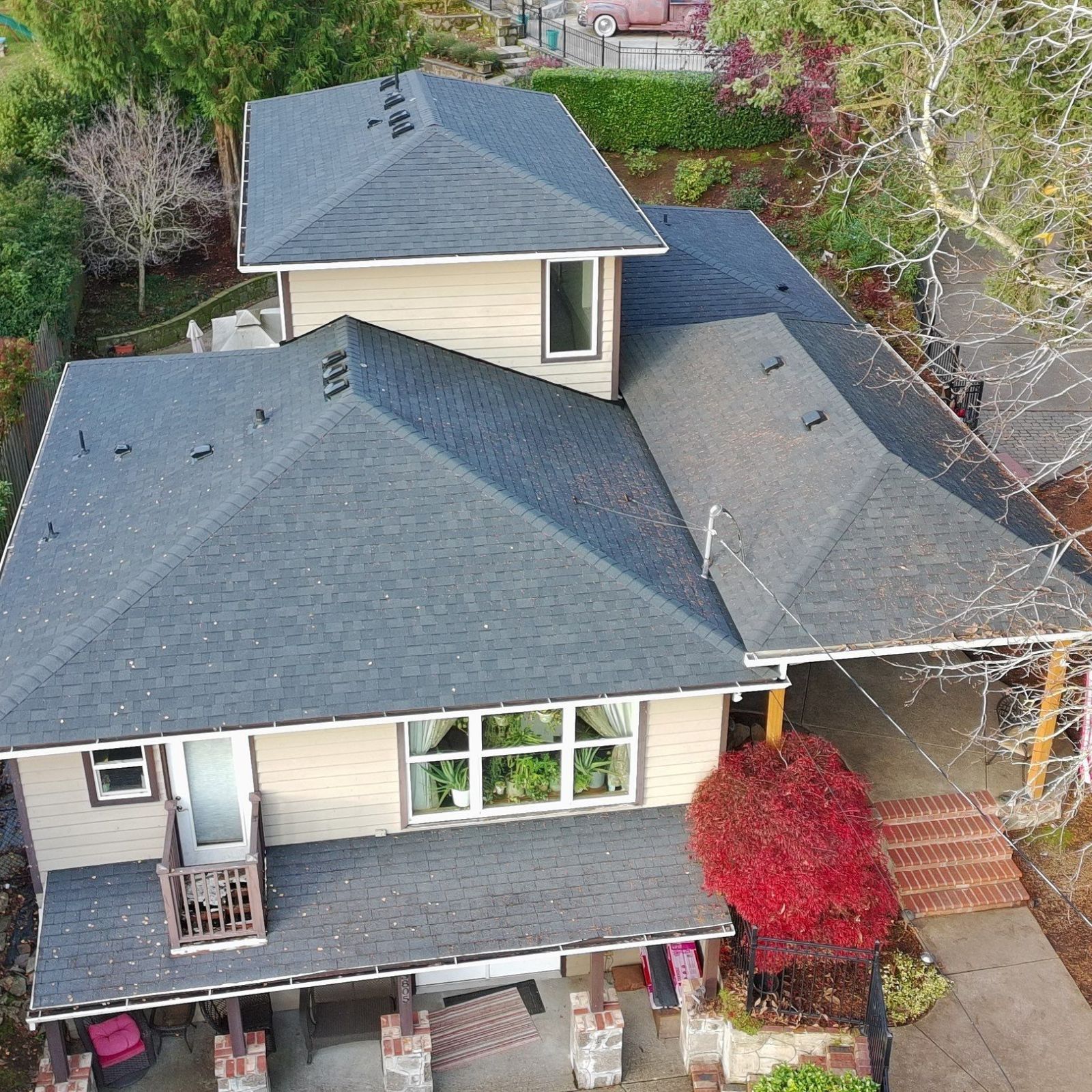 An aerial view of a dark shingled roof on a blue house, featuring chimneys, a skylight, and a small backyard patio.