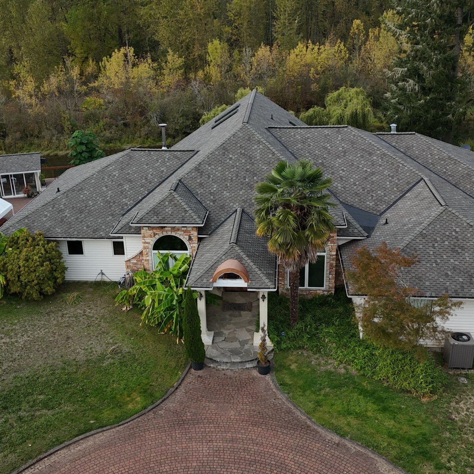 A high-angle view of a gray asphalt shingle roof with several vents, with trees and a house wall visible nearby.