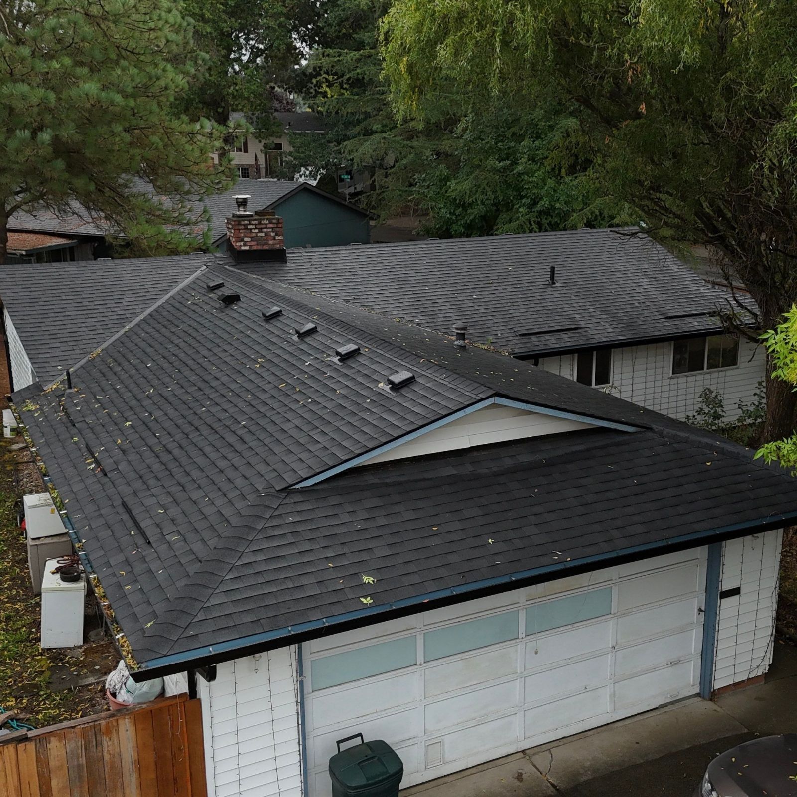 High-angle view of a dark shingled roof on a white house, showing vents, a chimney pipe, and a partially visible street.