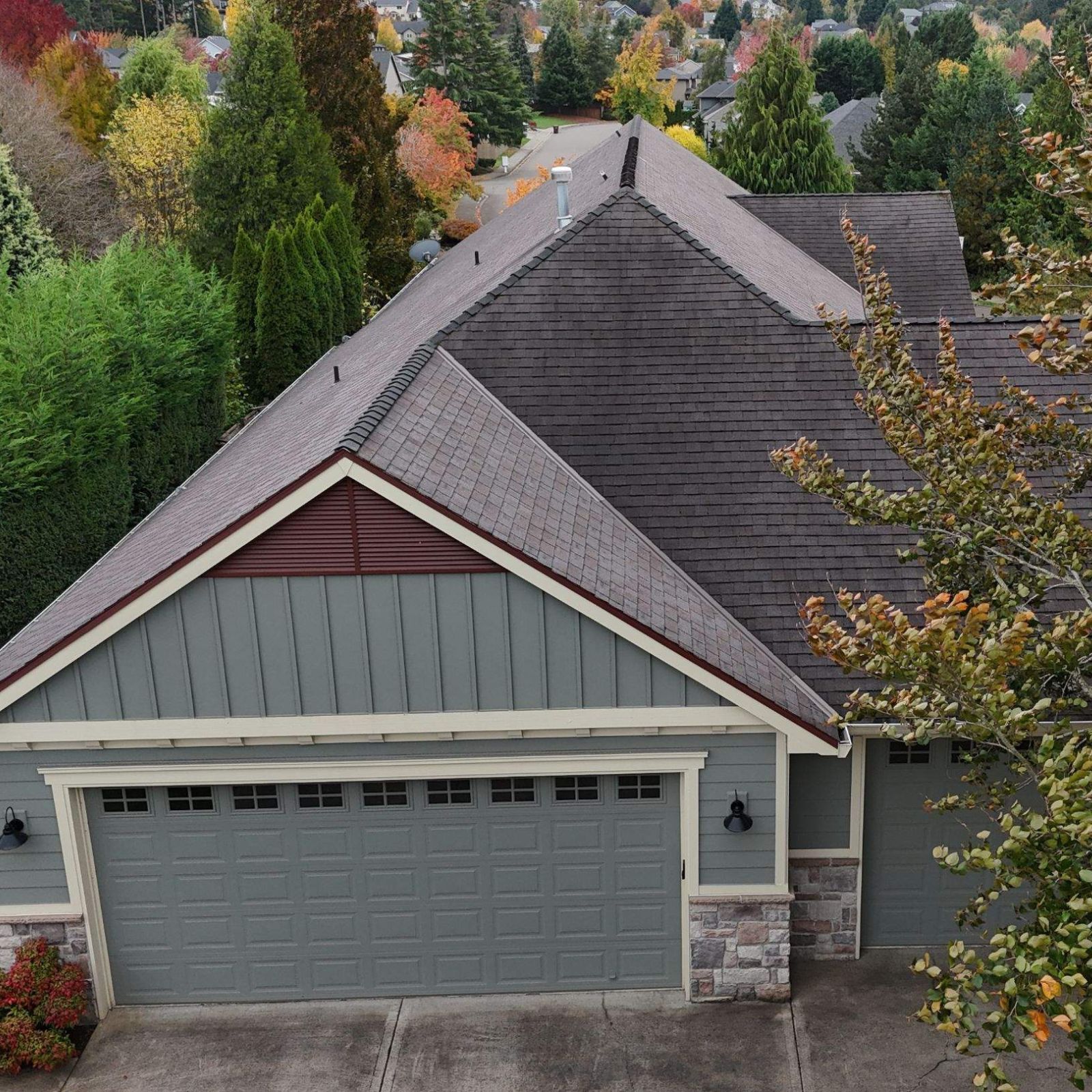 A high-angle view of a grey house garage with a dark shingled roof, surrounded by autumn trees.