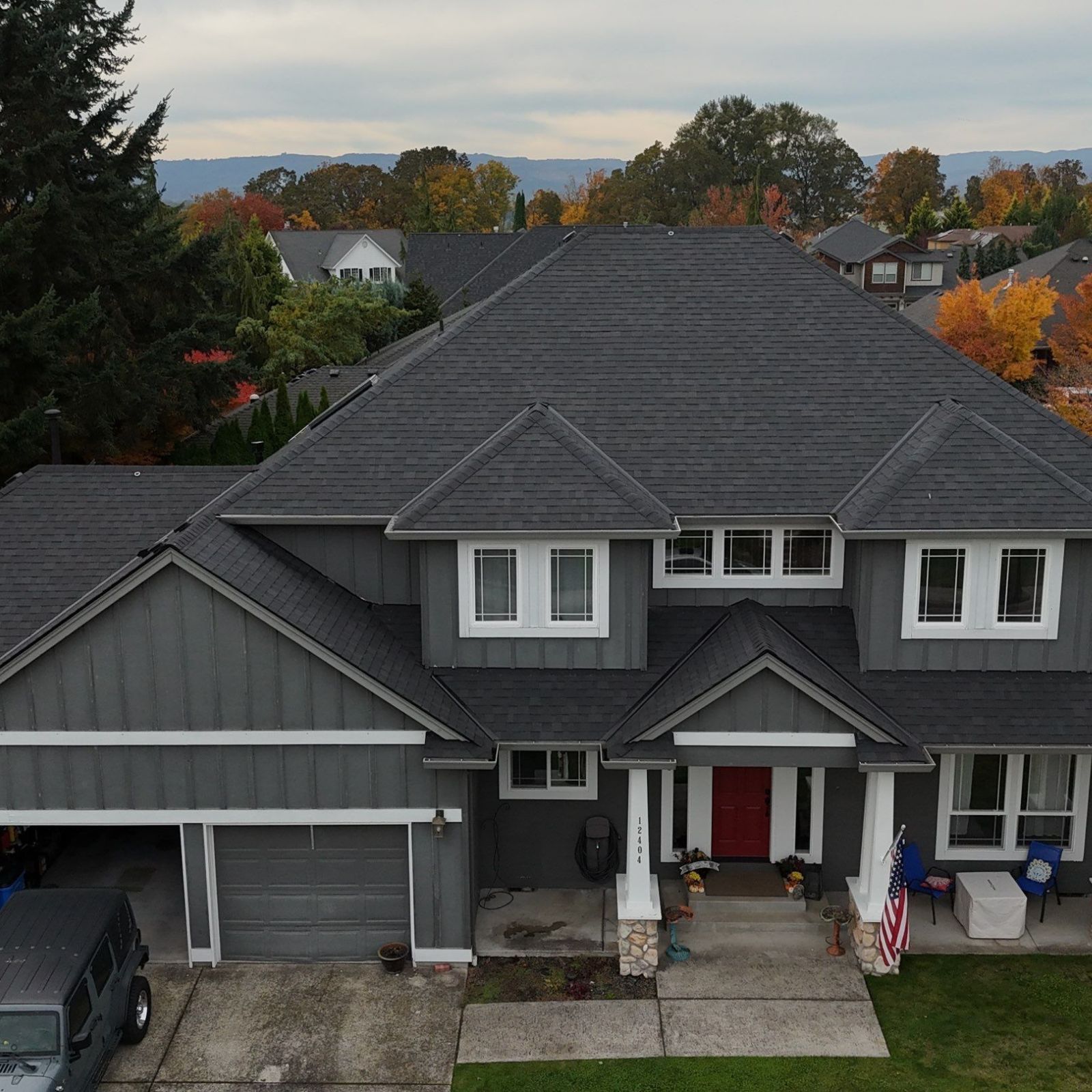 A two-story gray house with a dark shingled roof, a red front door, and a paved driveway, viewed from an elevated angle.