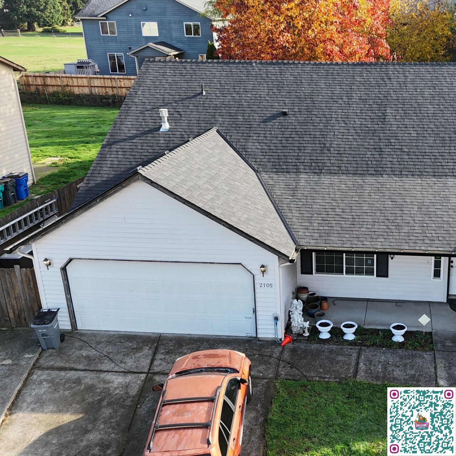 High-angle, aerial view of a tan two-story house with a grey shingled roof, featuring a front porch and red foliage.