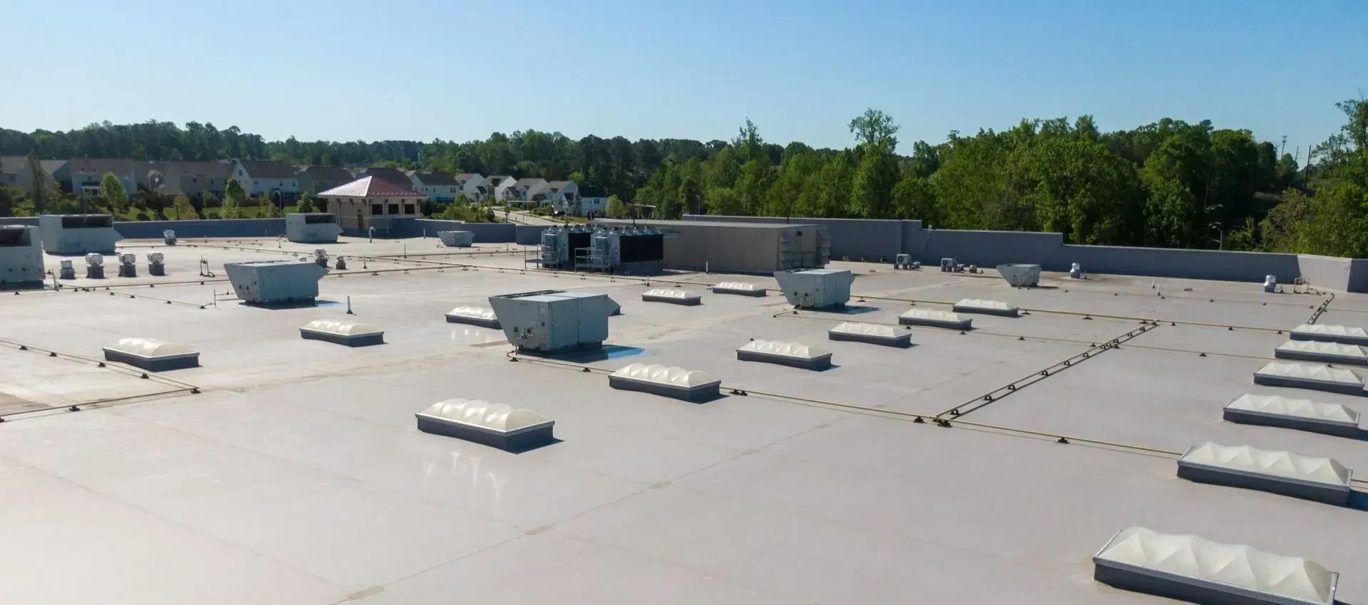A low-angle view of a flat roof with several air conditioning units and skylights; background includes trees and buildings.