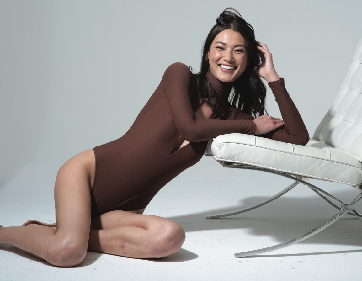 A woman in a brown bodysuit is kneeling next to a chair