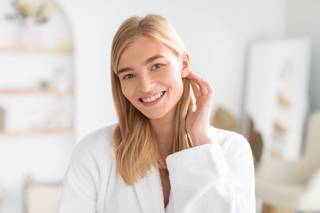 A woman in a white robe is smiling and touching her ear.