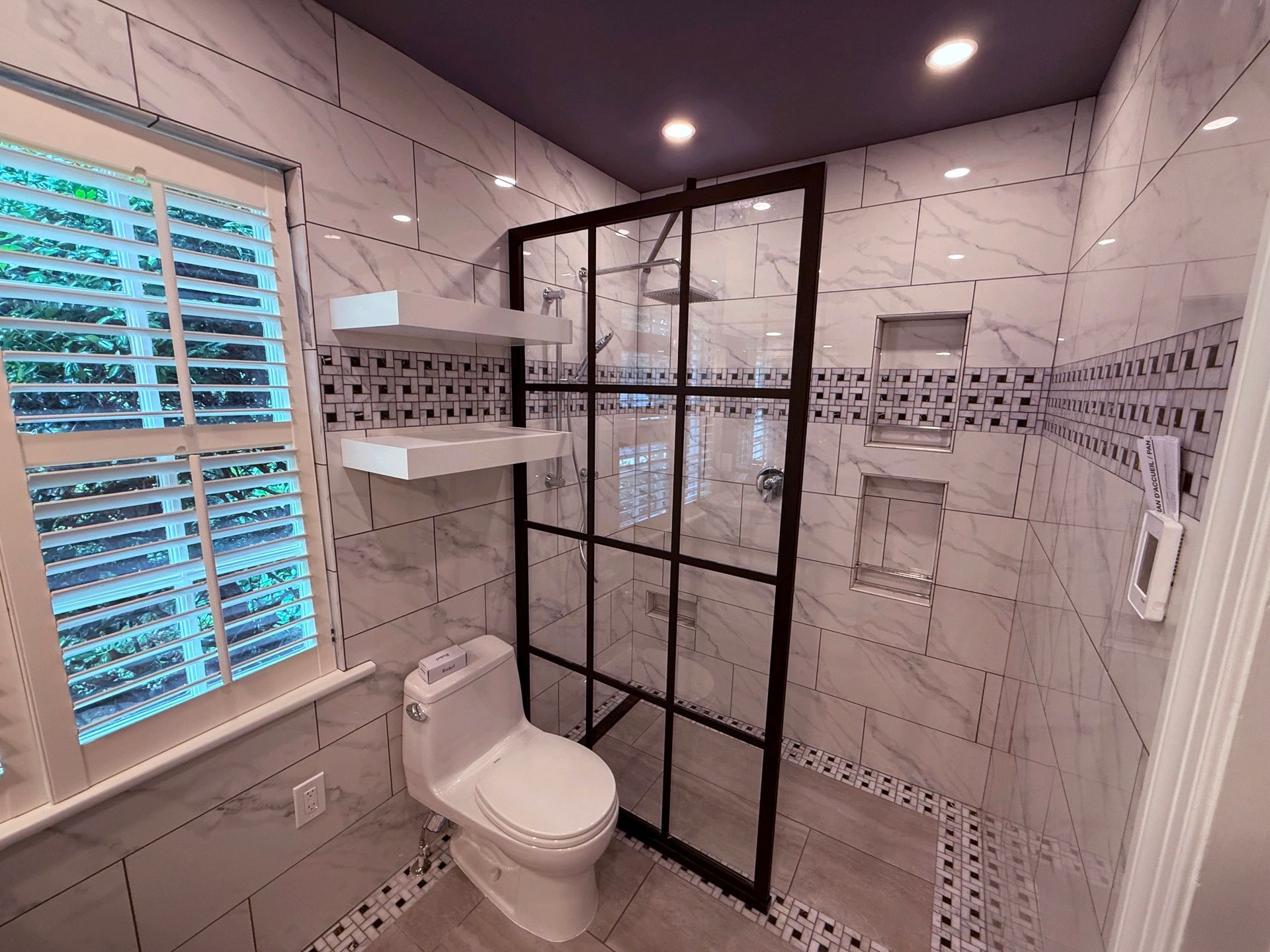 Bathroom with white marble tile, black shower enclosure, toilet, and window with shutters.