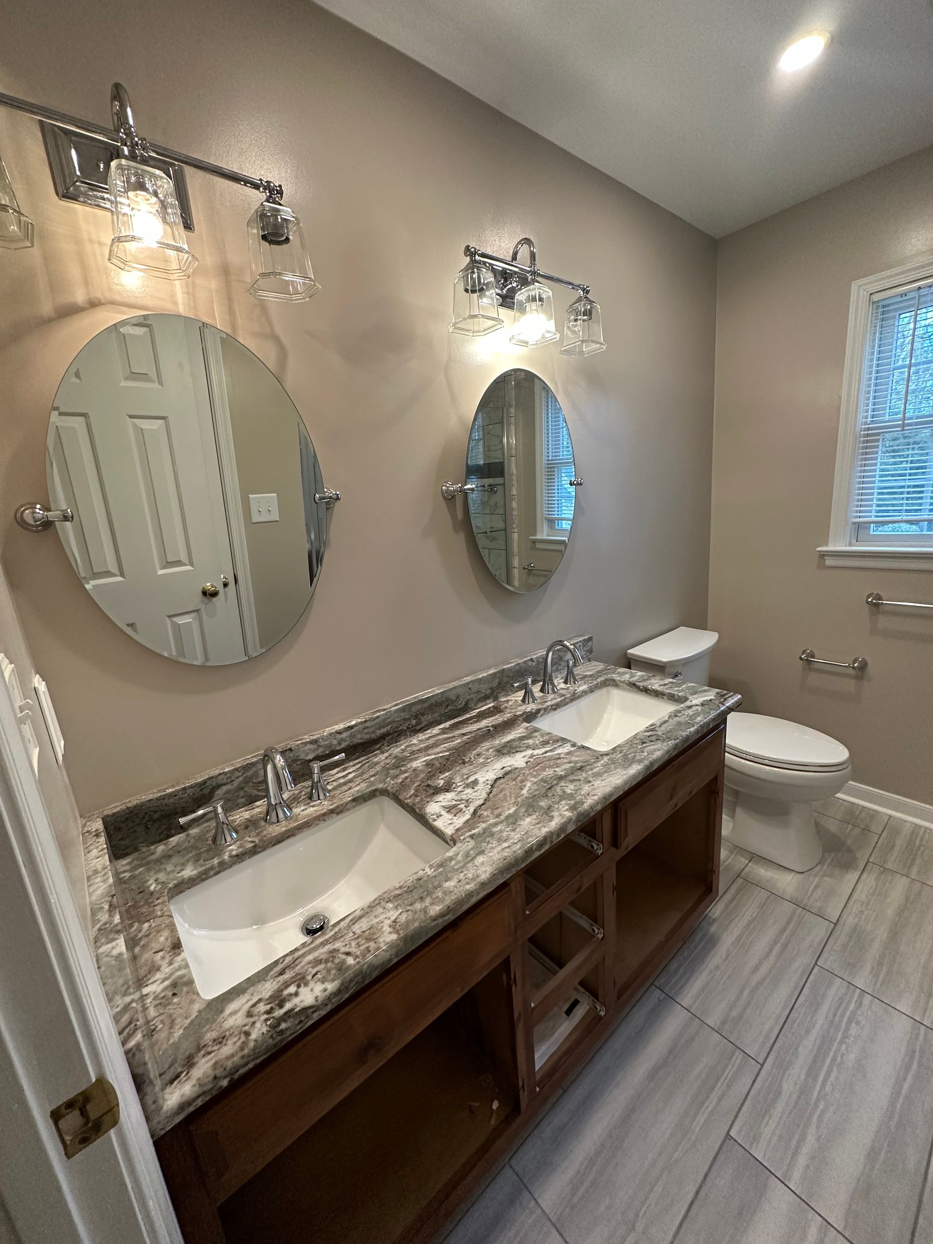 Bathroom with double sink vanity, oval mirrors, two sconces, toilet, and window.
