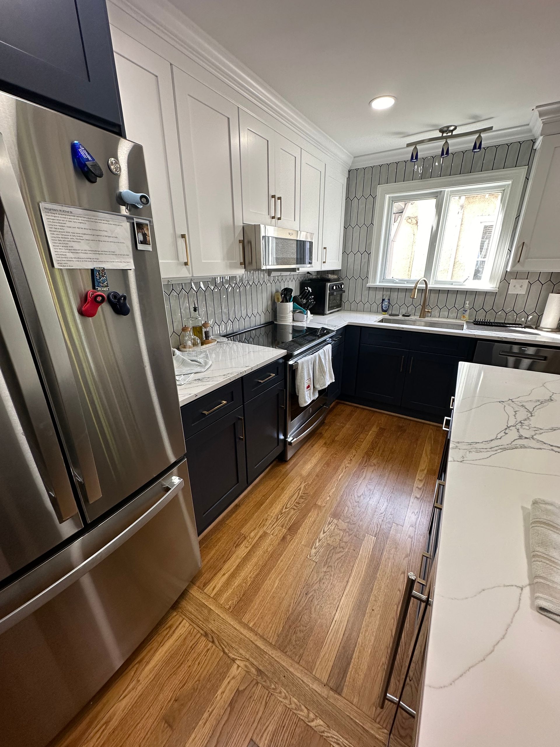 Kitchen with white and dark cabinets, stainless steel fridge, wood floor, and patterned backsplash.