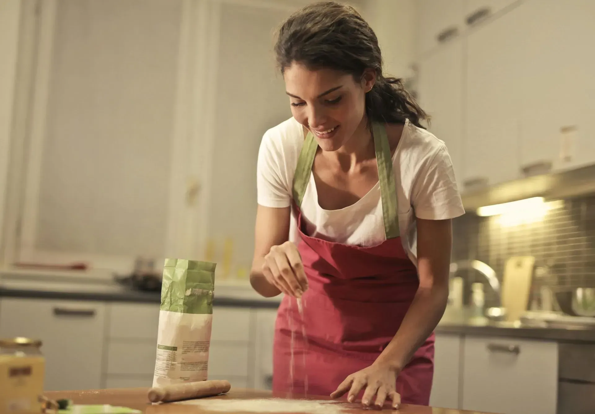 Woman in apron sprinkling flour in a kitchen.