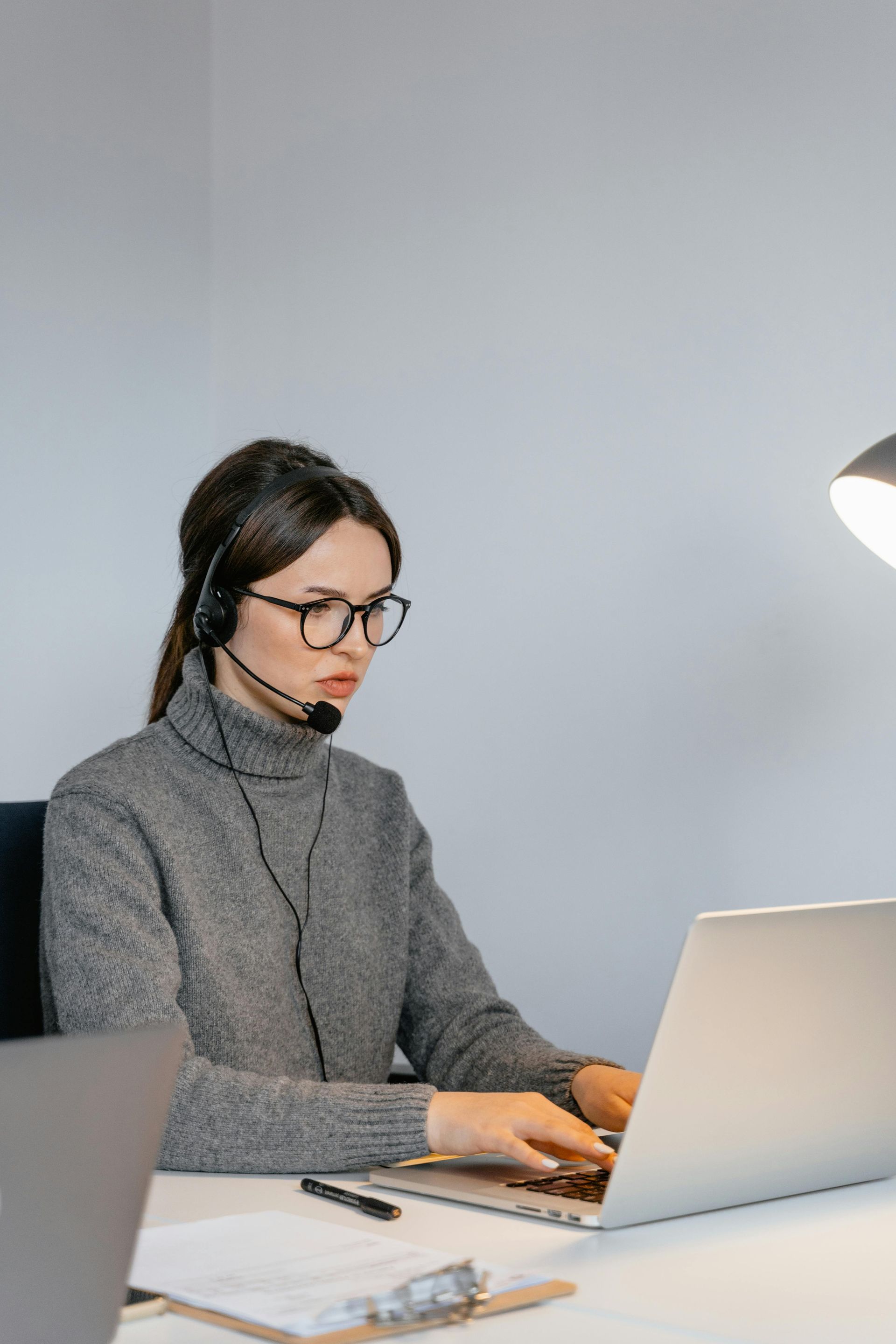 Woman wearing headset and glasses, working on a laptop at a desk with a lamp.