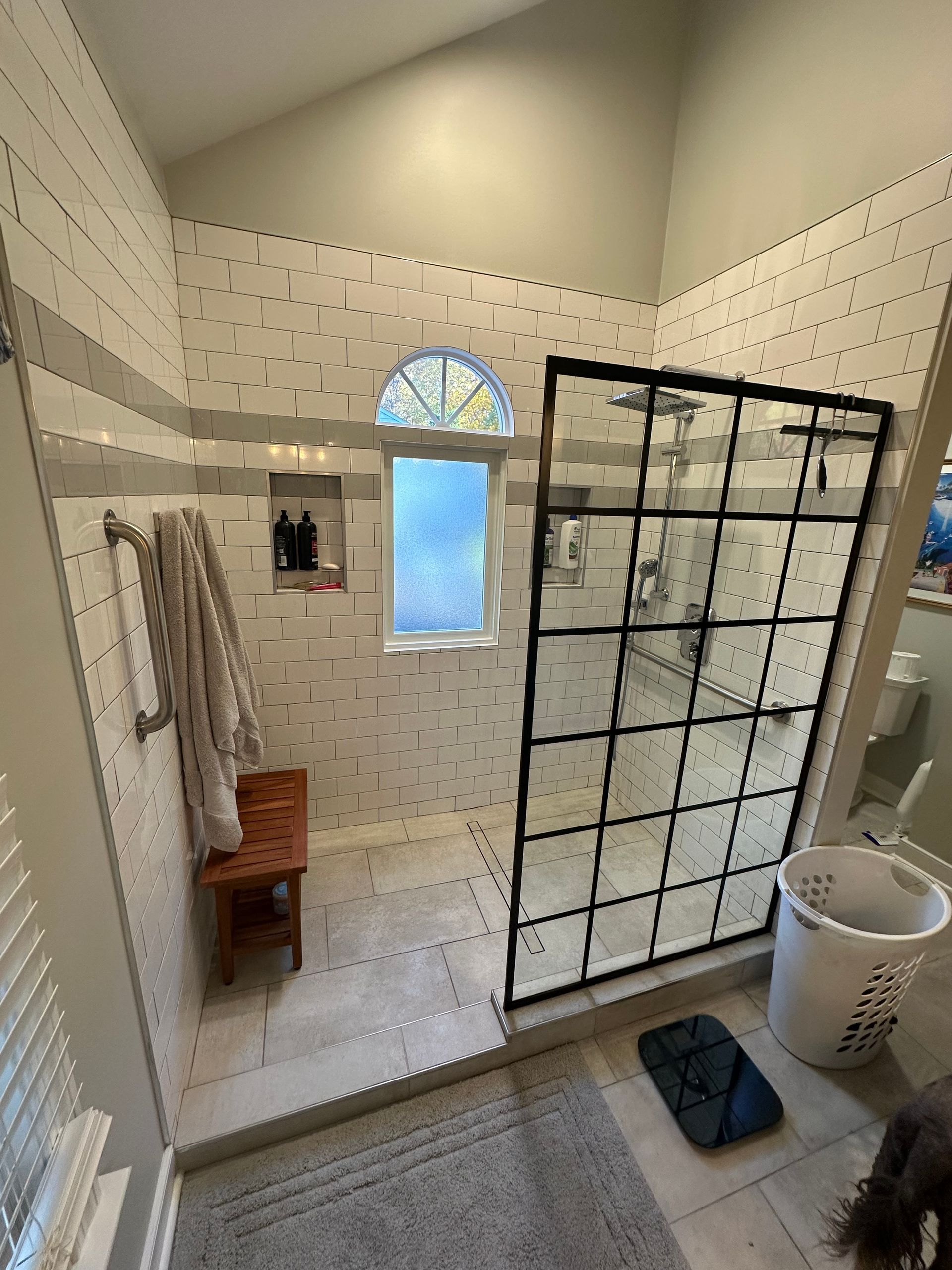 Bathroom with a tiled shower, black-framed glass door, small window, and a wooden bench.