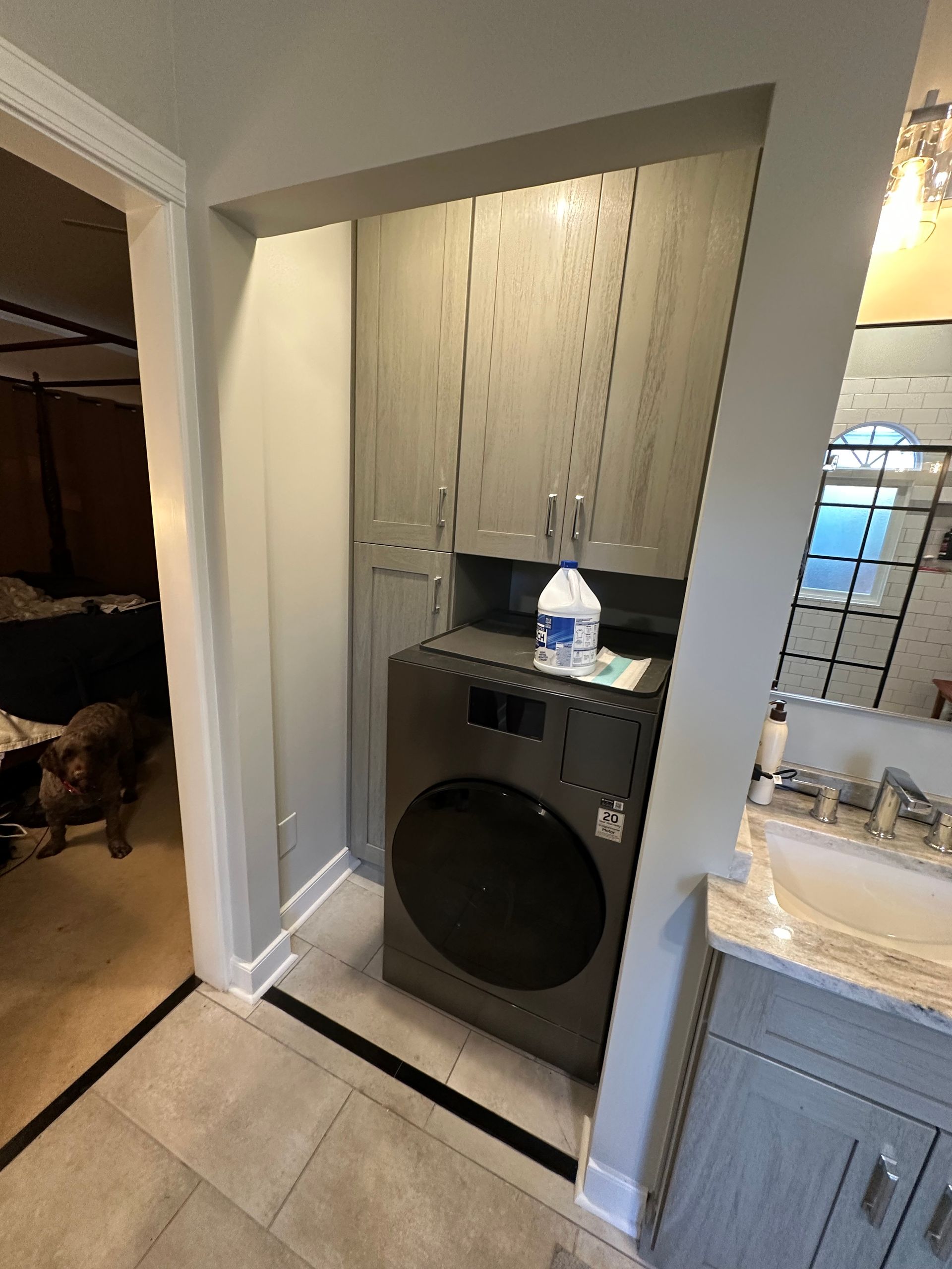 Laundry area in a bathroom with washer, cabinets, and laundry detergent. Dog peeks from doorway.