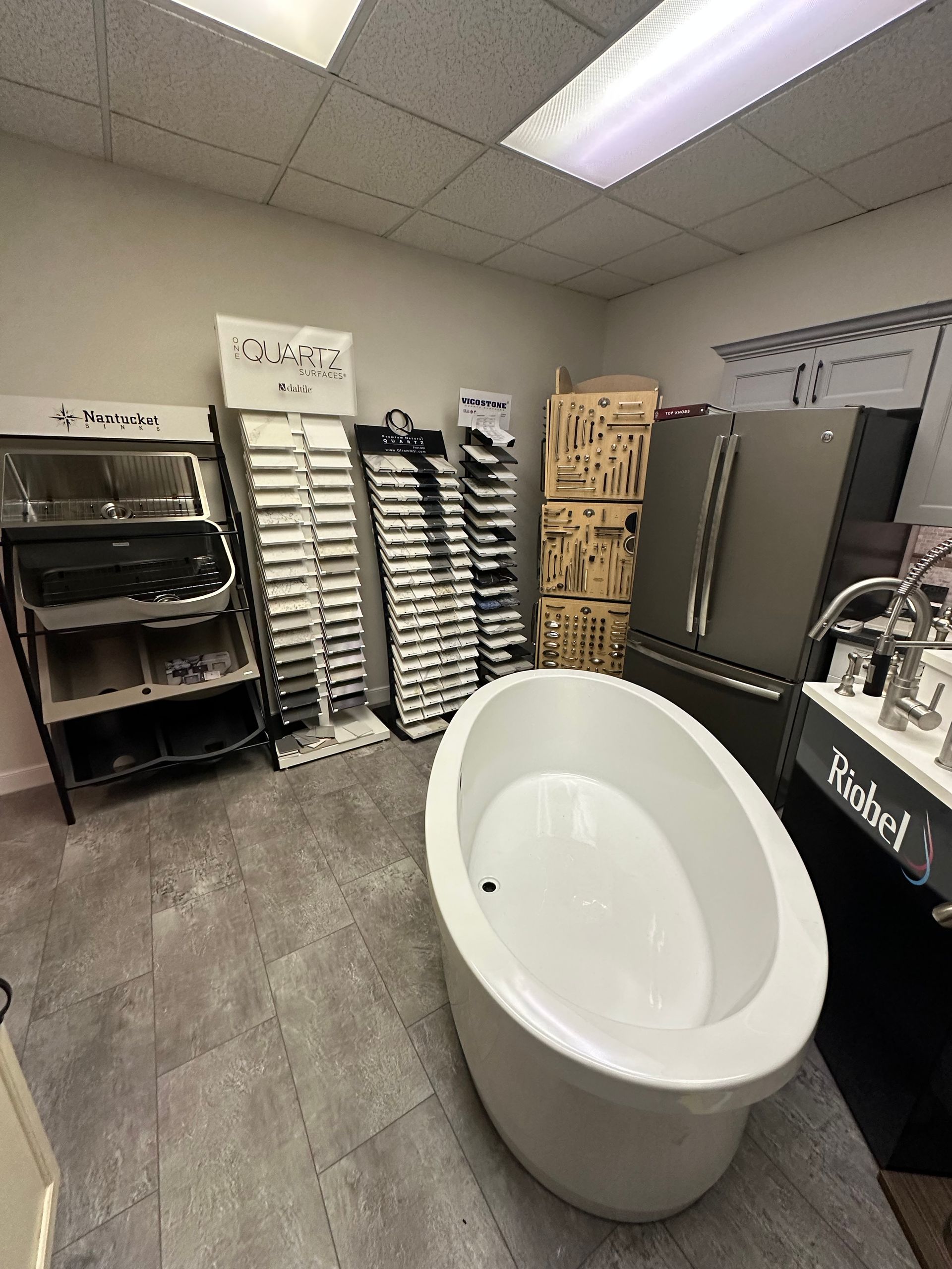 Bathroom showroom with a bathtub, refrigerator, and displays of various fixtures. Gray tile floor.