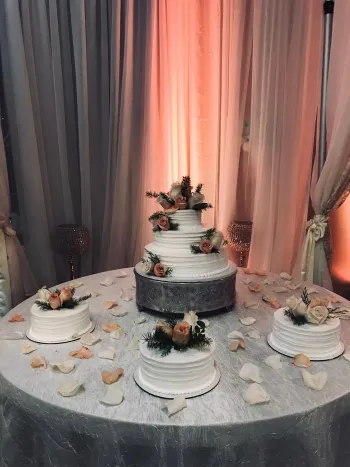 Elegant white tiered cake display on a silver table with small matching cakes and floral decor in front of draped curtains
