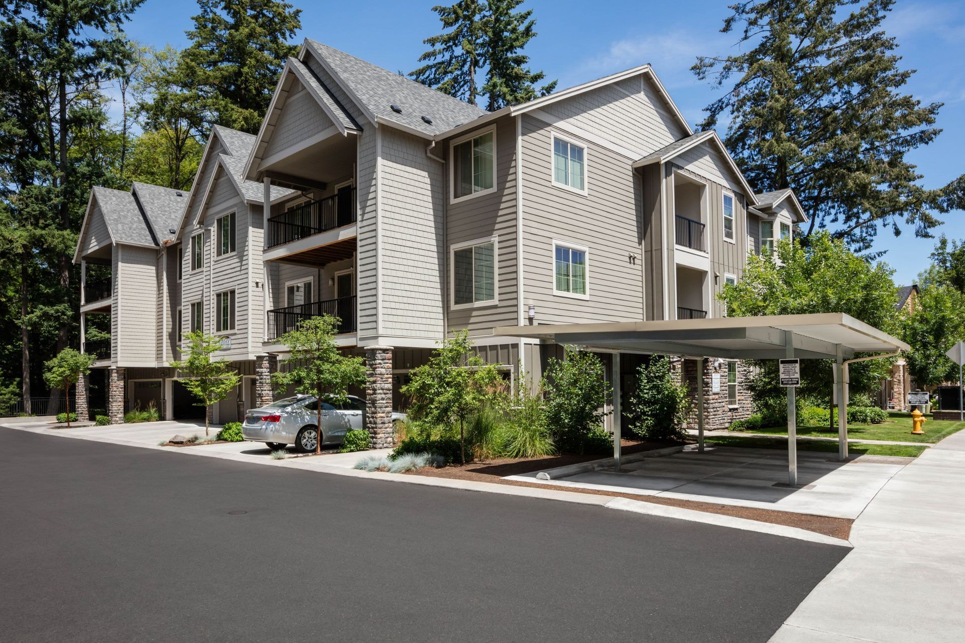 Exterior view of a modern, multi-story apartment building with a covered parking area and trees.