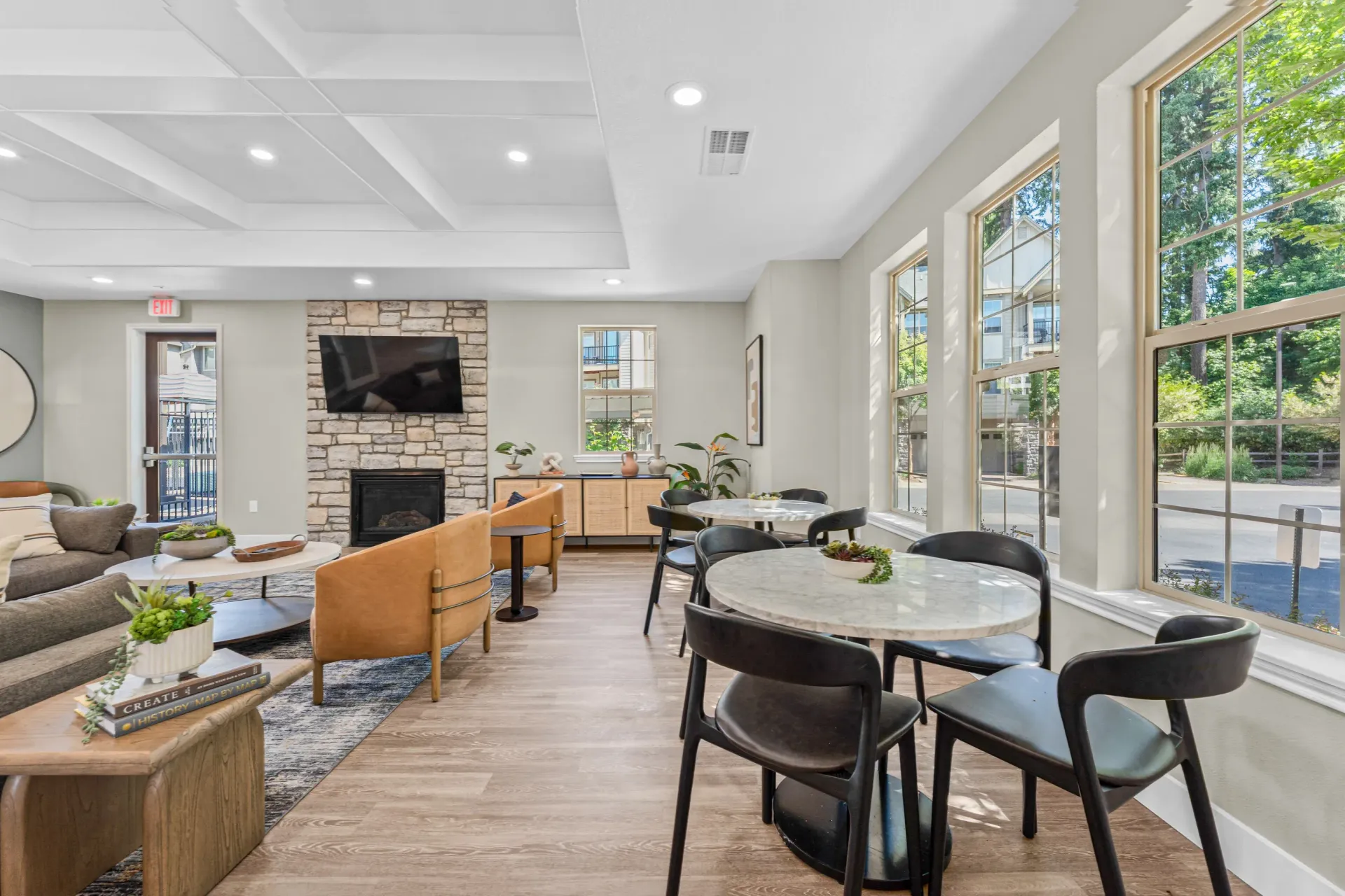 Bright communal lounge with sofas, round tables, a stone fireplace, and large windows.