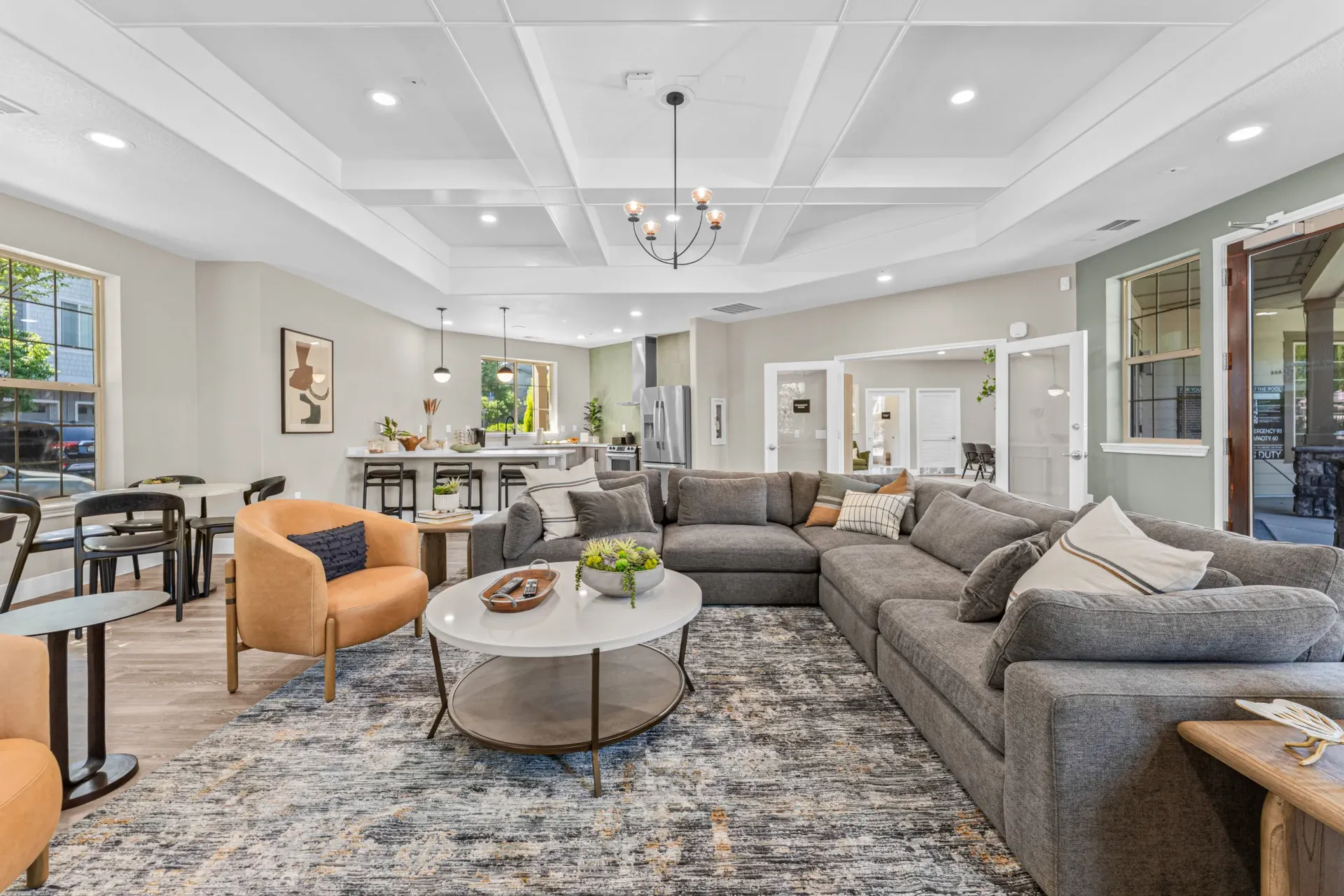 Bright communal lounge with gray sectional sofa, orange chairs, coffee table, and a kitchen area.