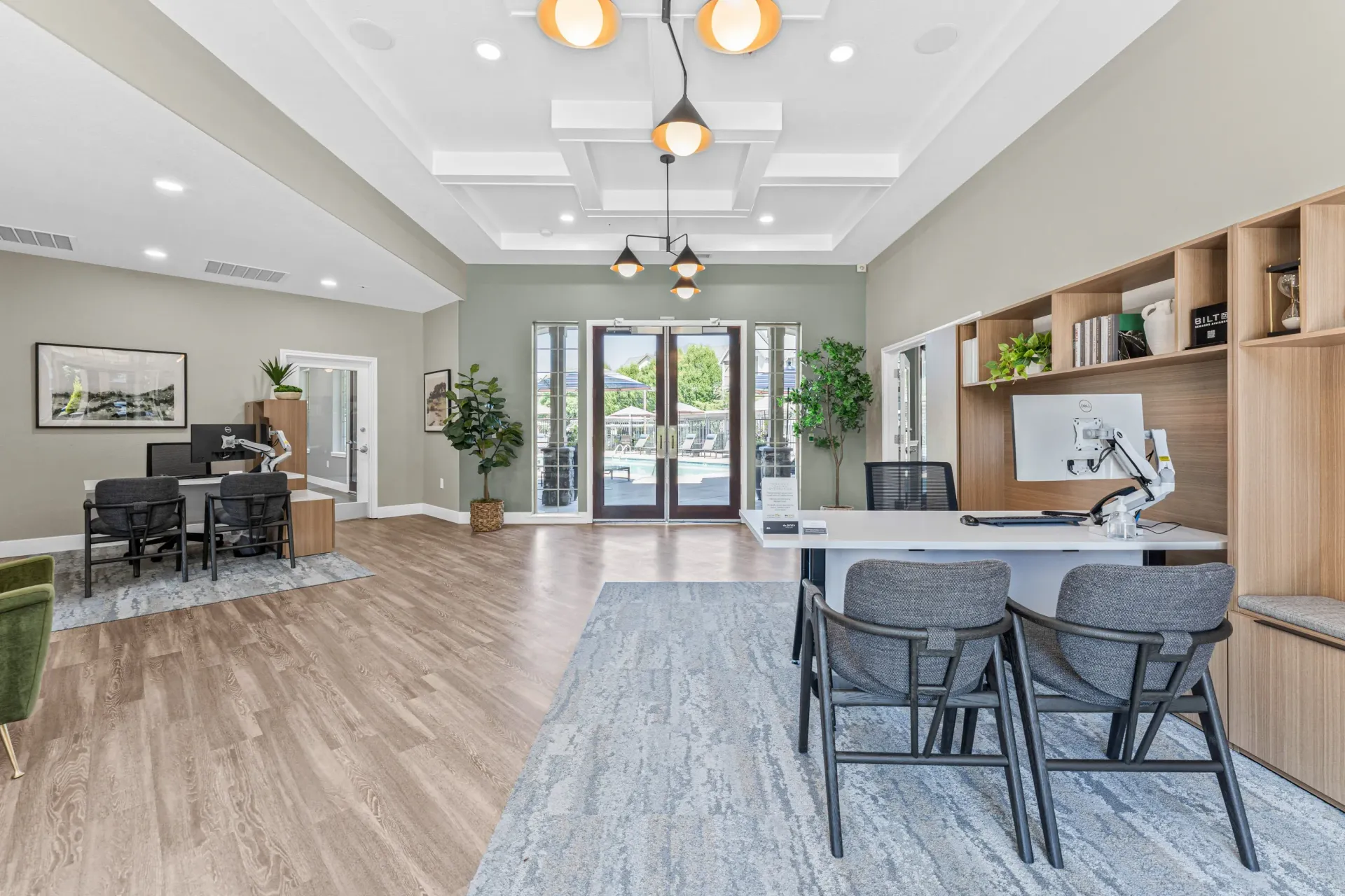 Modern apartment community lobby with a front desk, seating area, shelving, and glass doors to outdoor amenities.