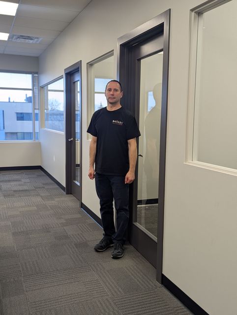 A person standing in an office hallway with patterned carpet and glass-paneled office doors.