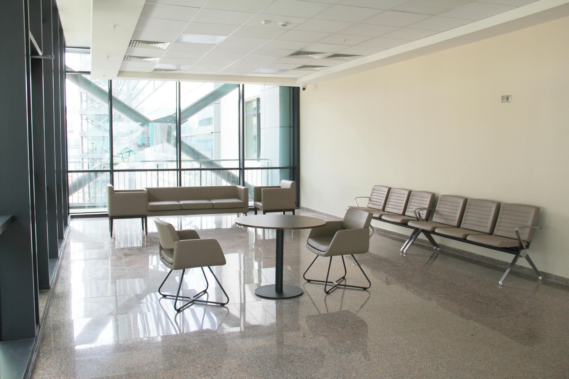 A waiting area with modern beige chairs, a circular table, and a long couch in a bright room with large glass windows.