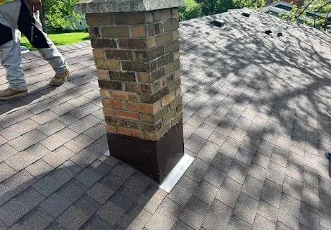 Brick chimney with dark metal flashing on a brown shingle roof, person standing.