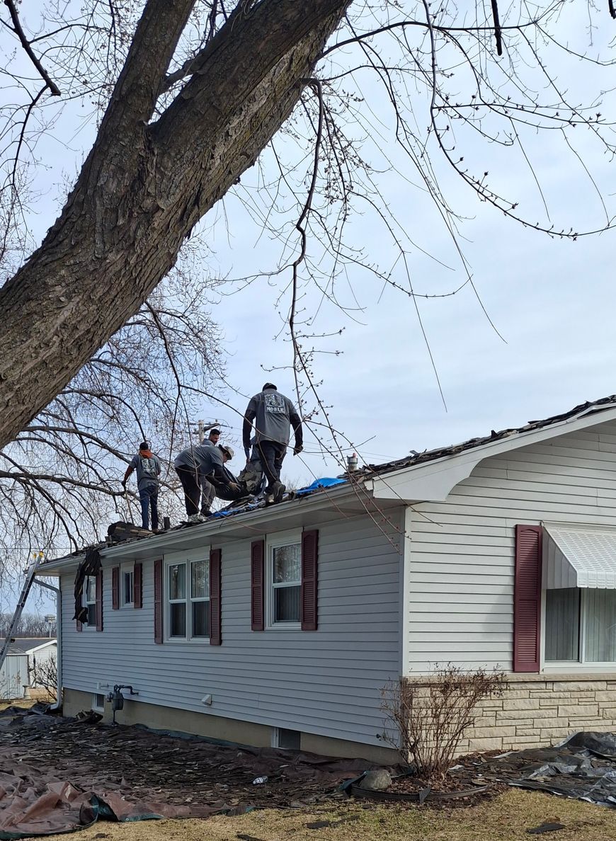 Three people on a roof repairing damage; white siding, brown shutters, tree in foreground.