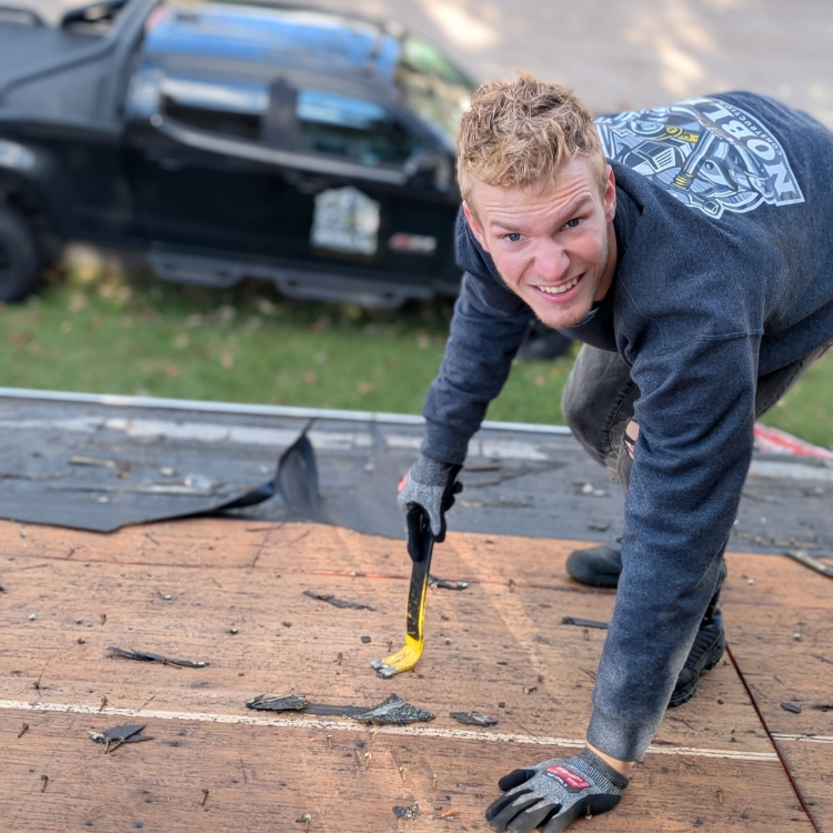 Blonde man removing roofing shingles with a pry bar. Dark shirt and gloves. Car in background.