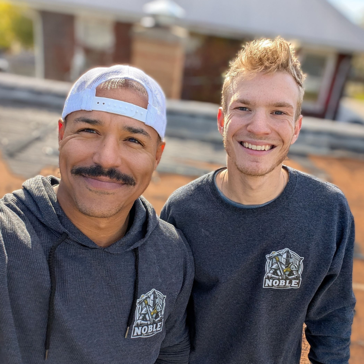 Two men smiling, wearing gray sweatshirts and a white cap; outdoors.