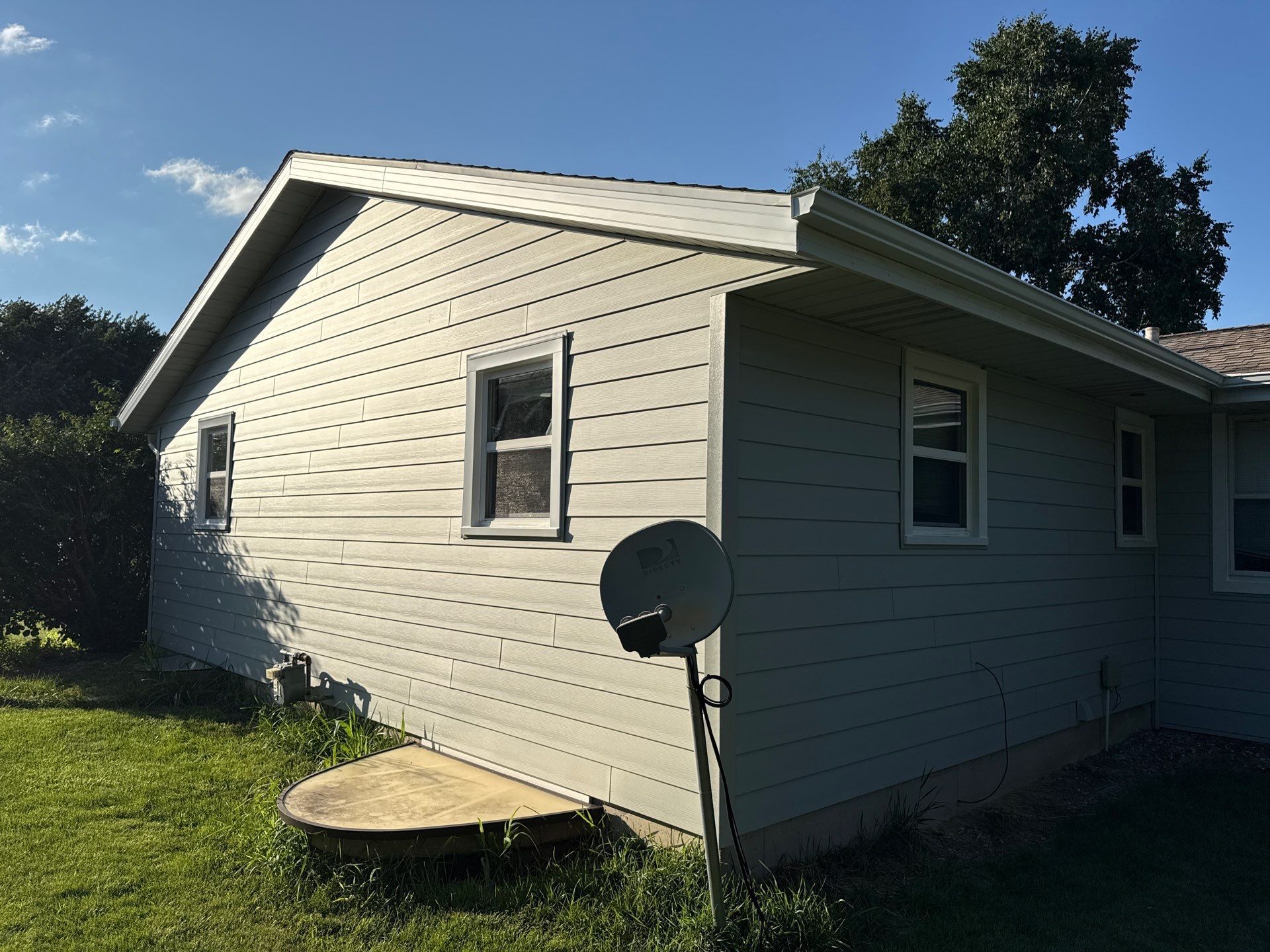 A light green house with gray roof, windows, and satellite dish. Sunny day, grassy yard.