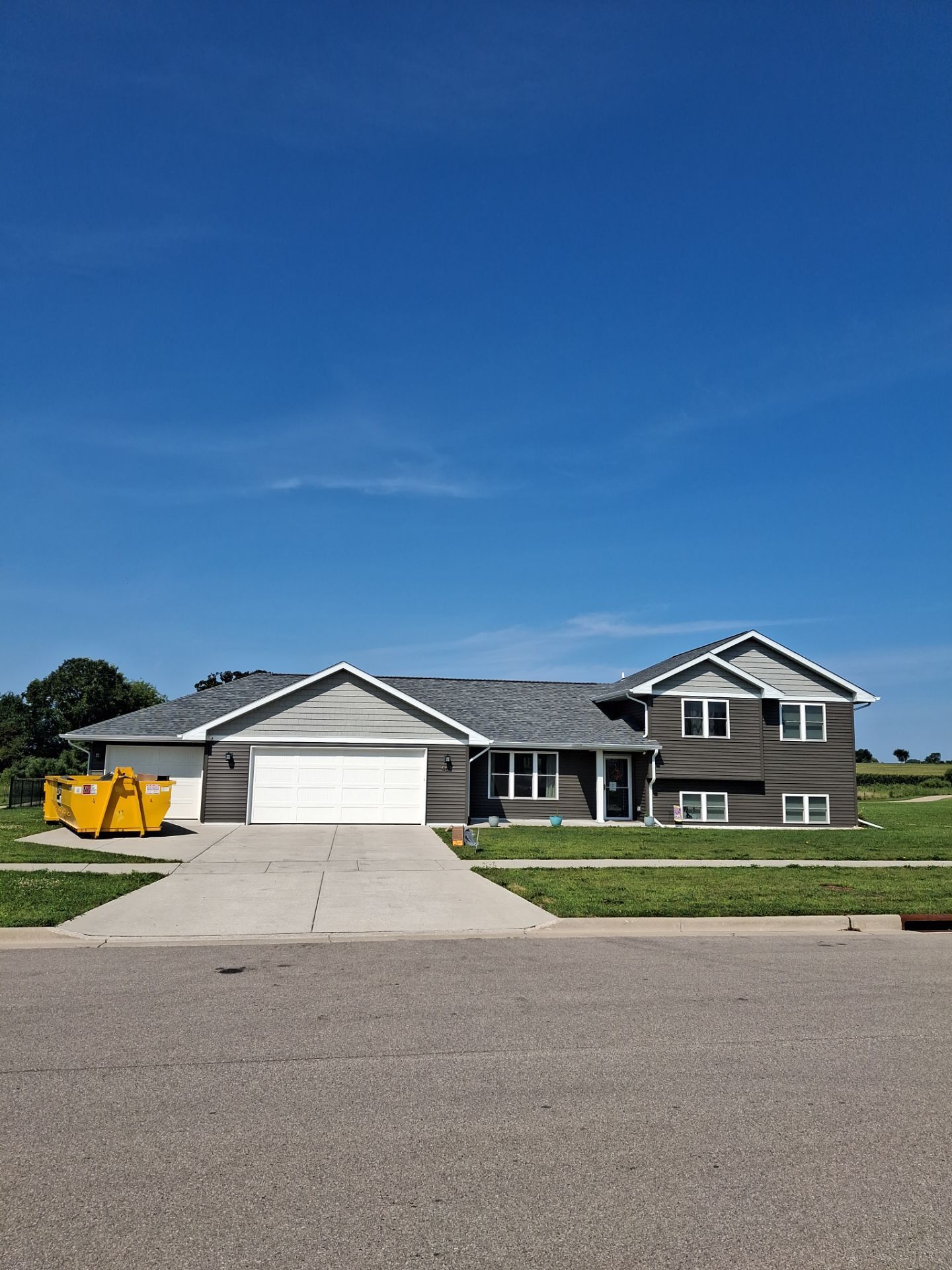 Gray house with white garage door, blue sky. Concrete driveway and yellow dumpster visible.