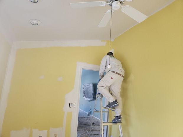 A person on a ladder is installing a light fixture in a room with yellow walls.