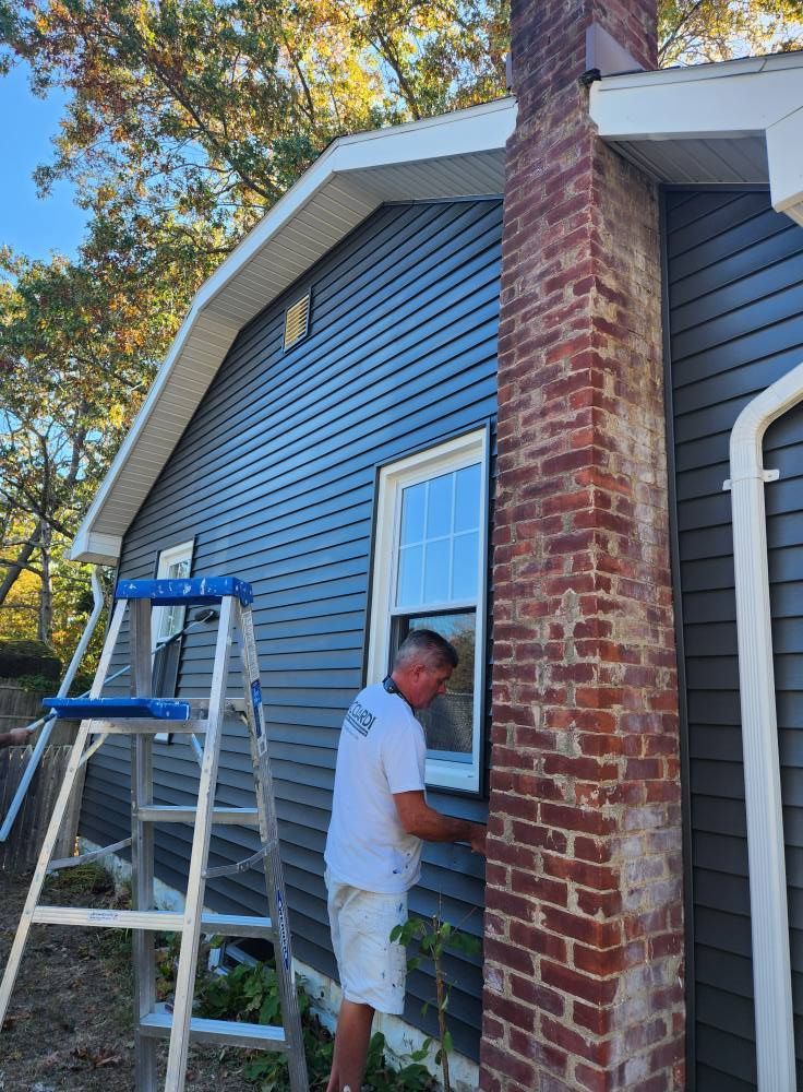 Man on a ladder, installing a window frame, next to a brick chimney and blue siding.