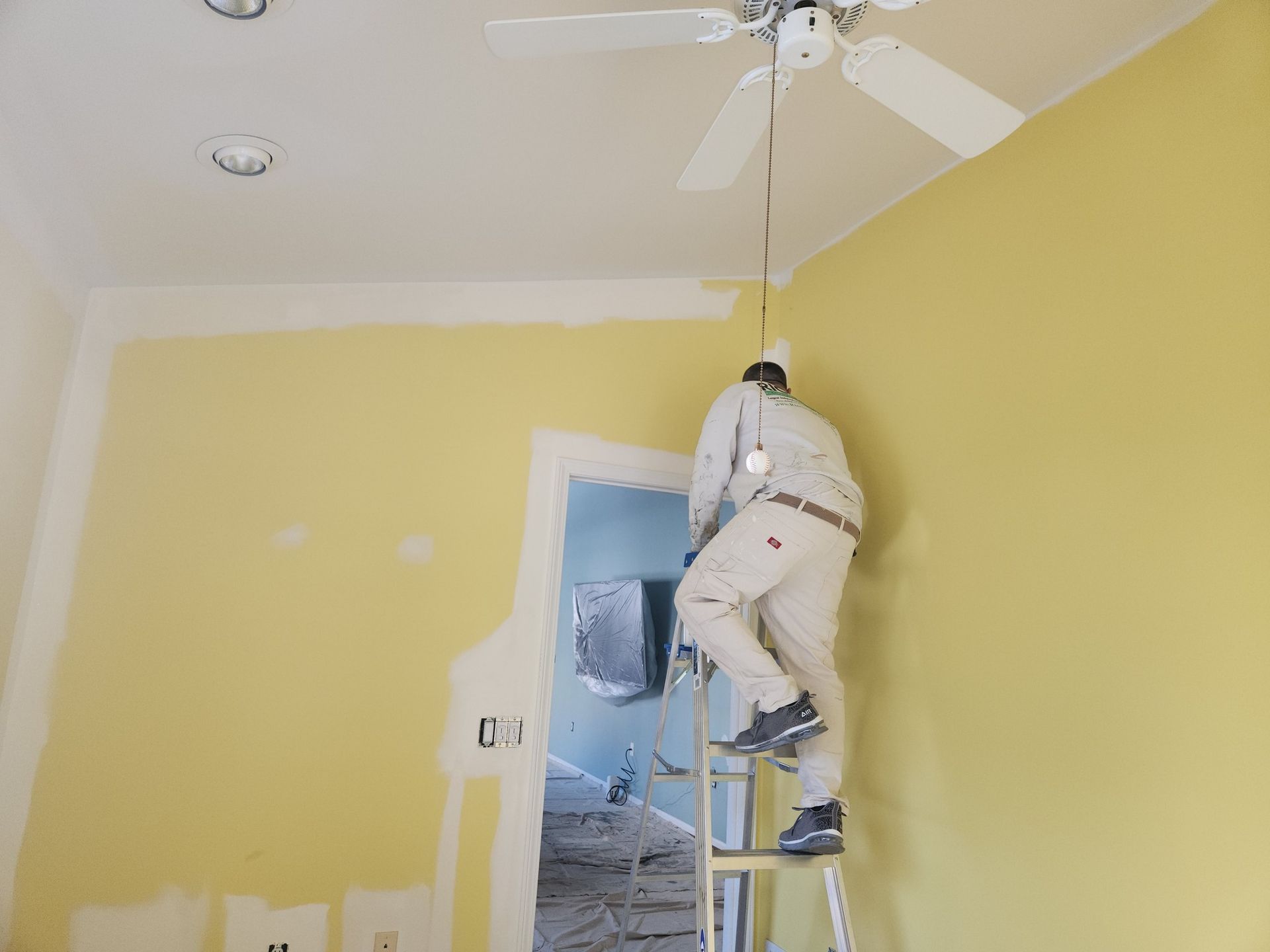 Person on a ladder, working on a light fixture in a room with yellow walls.
