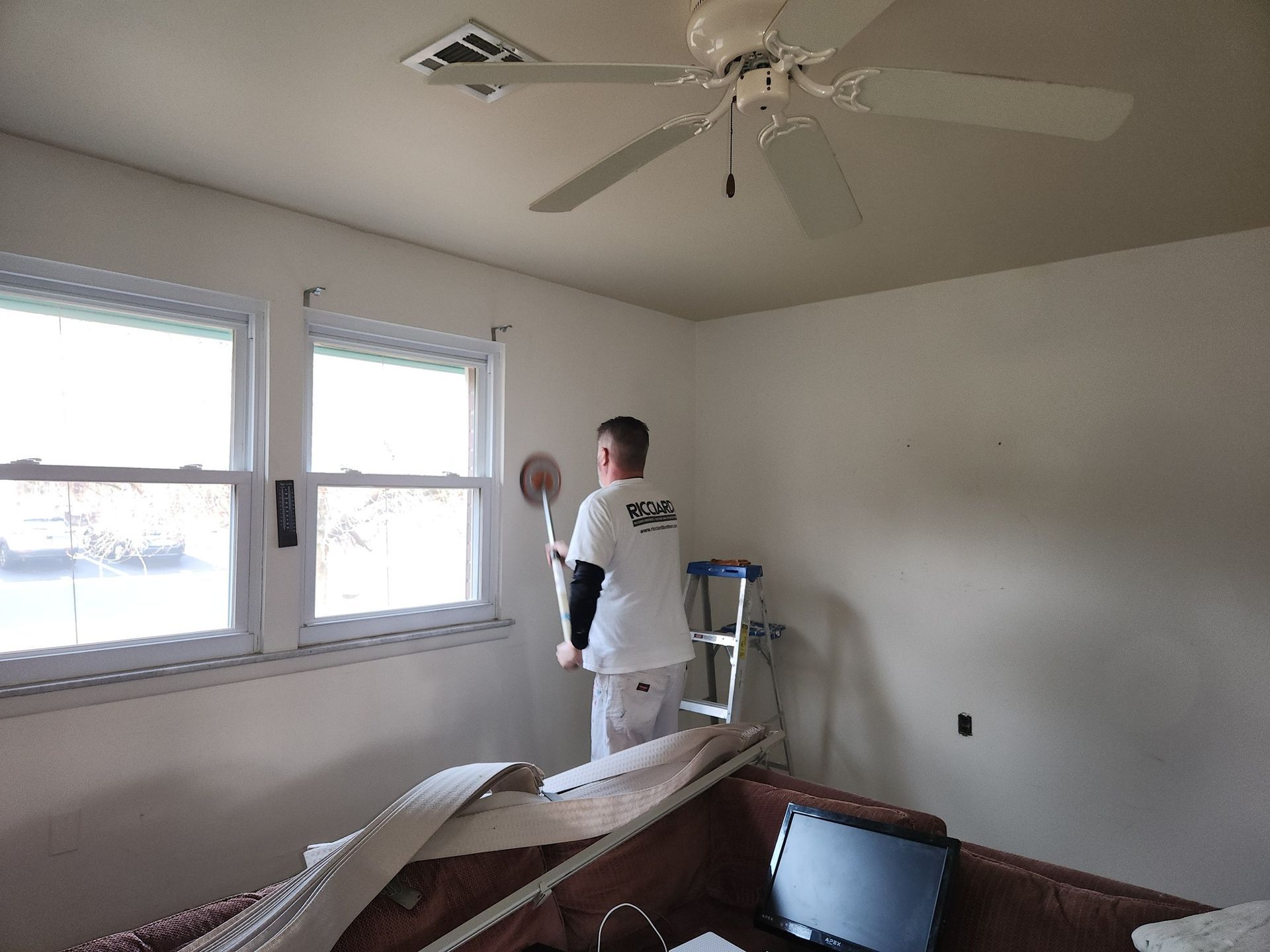Person painting a white wall indoors with a roller; a sofa and windows are also present.