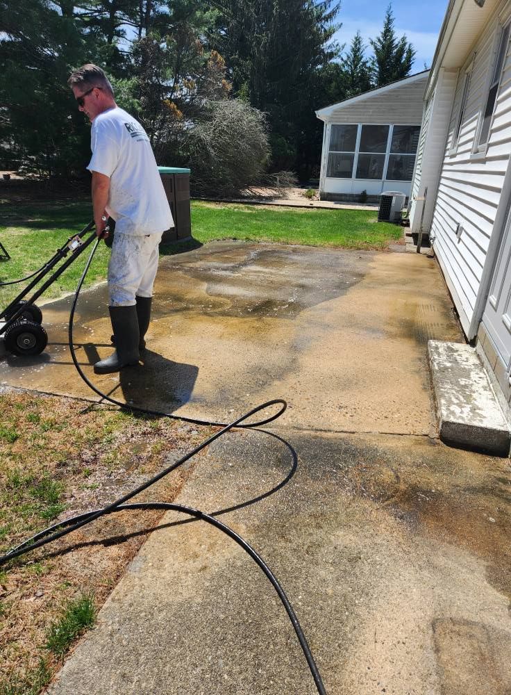 Person power washing a concrete patio next to a white house; sunny day.