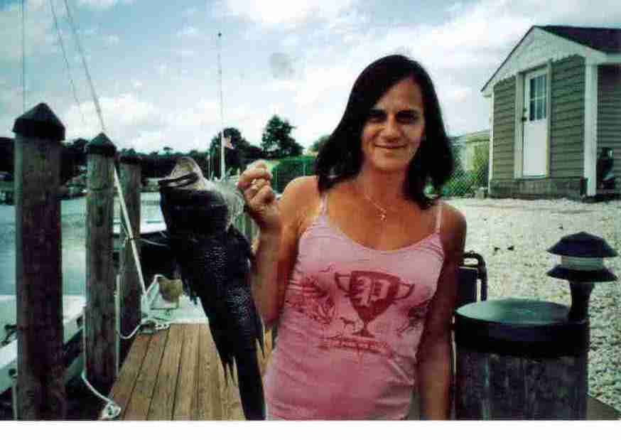 Woman on a dock holds a fish. She wears a pink tank top and smiles. Wooden dock, water, and building in background.