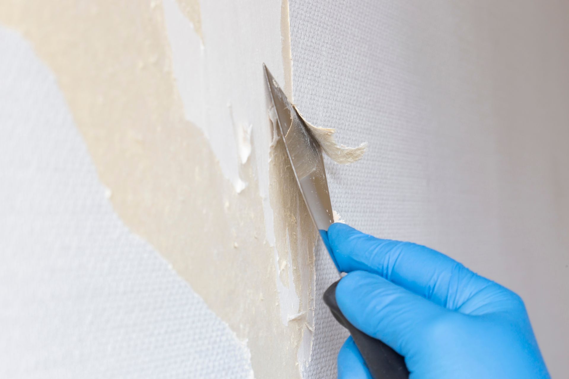 Gloved hand using a scraper to remove old wallpaper from a white wall.