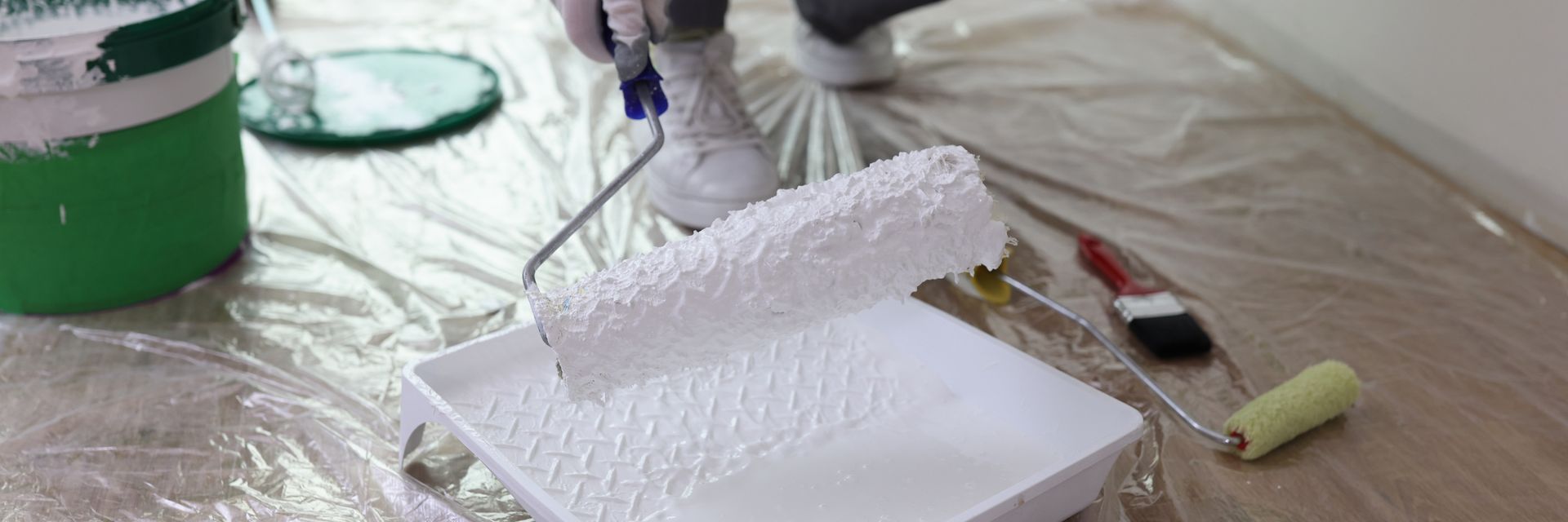 A person using a paint roller in a tray of white paint, with tools and a paint bucket, on a drop cloth.