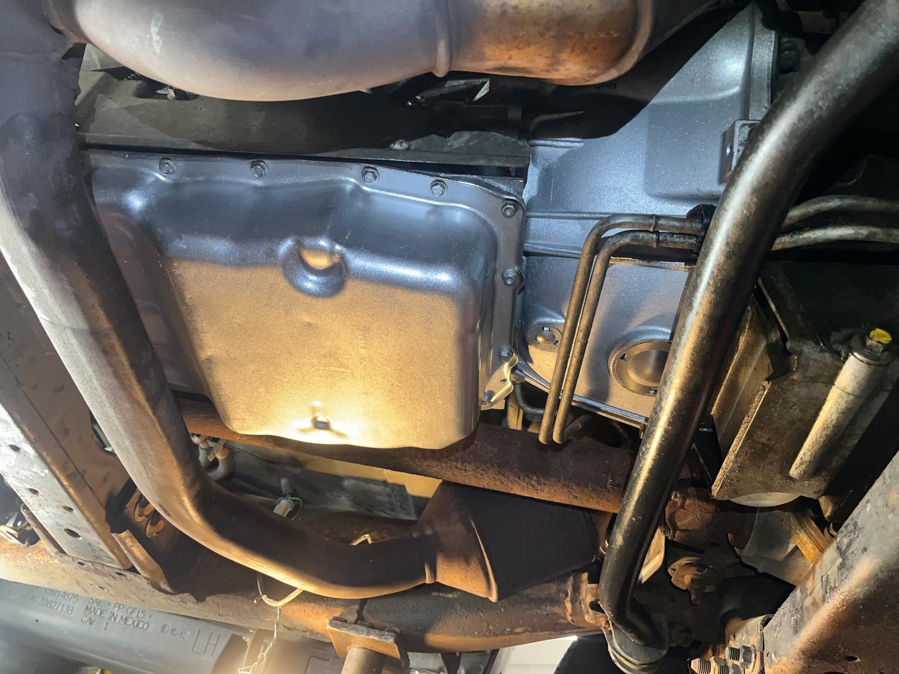 Undercarriage view of a vehicle; silver oil pan and transmission, exhaust pipes, and rusty frame are visible.