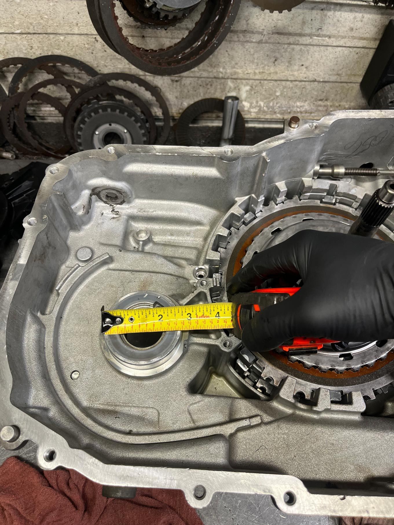 Gloved hand measures a transmission case with a tape measure. Parts and tools scattered on a workbench.