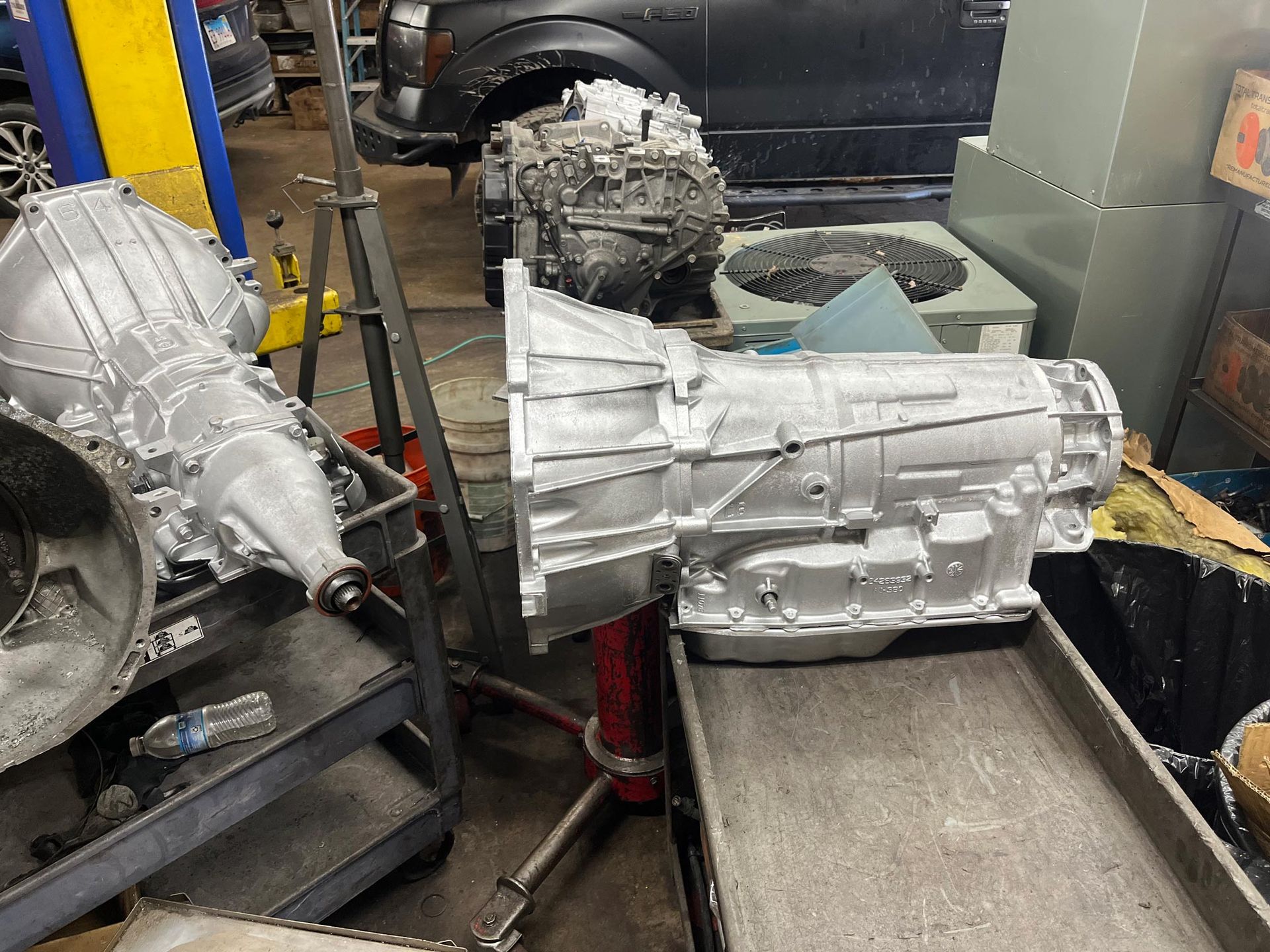 Three silver car transmissions in a mechanic's shop, one on a bench, others on stands.